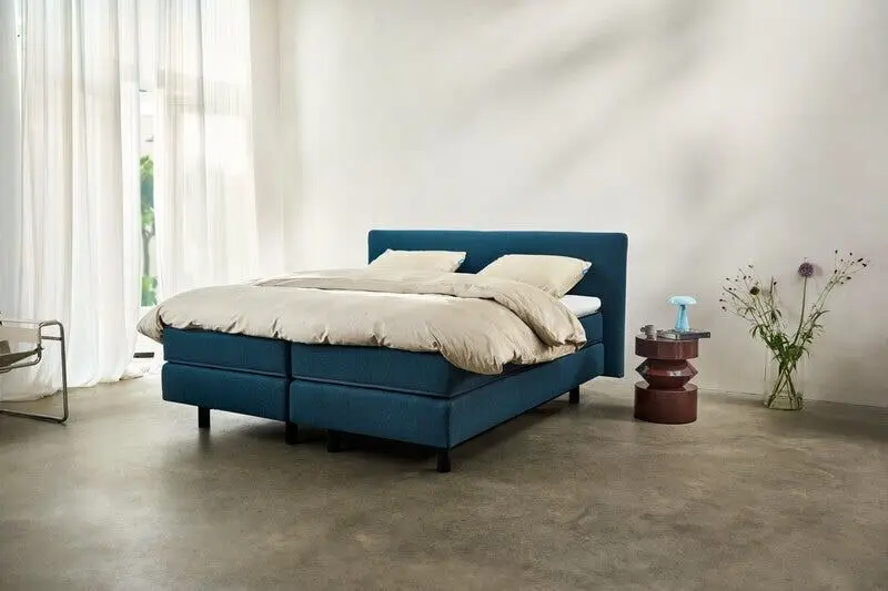 Petrolblauwe Auping-boxspring met lichte lakens op een betonvloer, met vitrages en een vaas bloemen