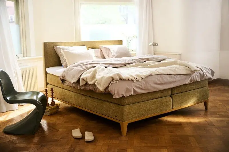Olijfgroene Auping-boxspring met linnen beddengoed en een wollen plaid, in zonlicht op een parketvloer
