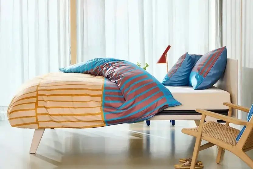 Wit Auping-bed met schuine houten poten en een gestreept dekbed voor lange vitrages, met een rieten stoel