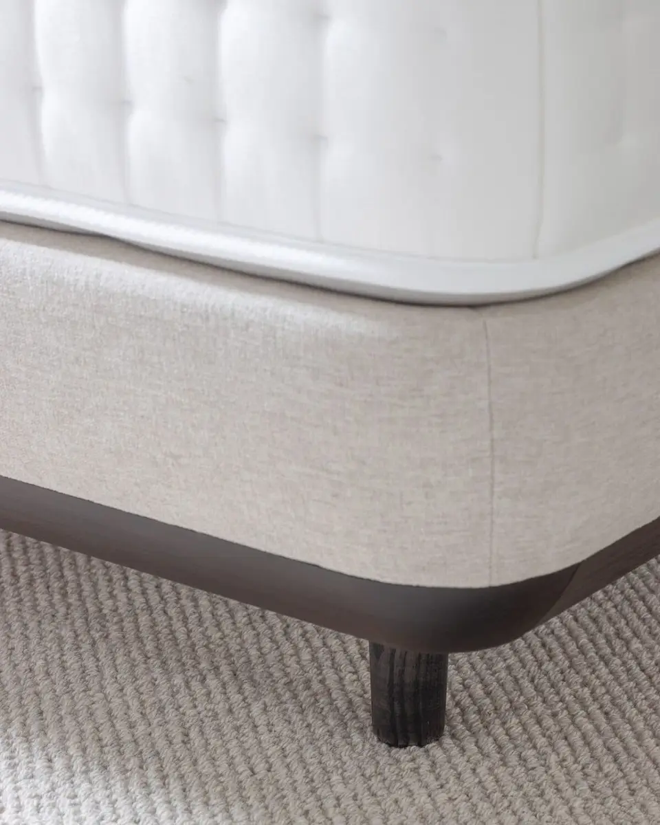 Hoek van een handgetufte matras op een linnen boxspring met een donkere plint, op een wollen kleed