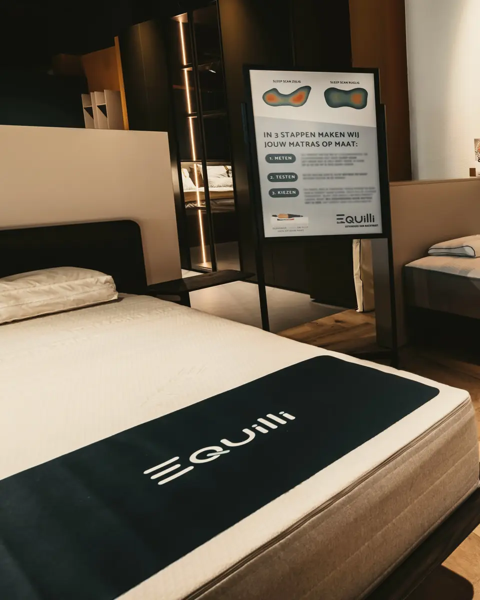 Equilli in onze showroom: een bord dat in drie stappen uitlegt hoe uw matras op maat wordt gemaakt, met twee drukbeelden van de Sleep Scan, naast een bed met een Equilli-matras