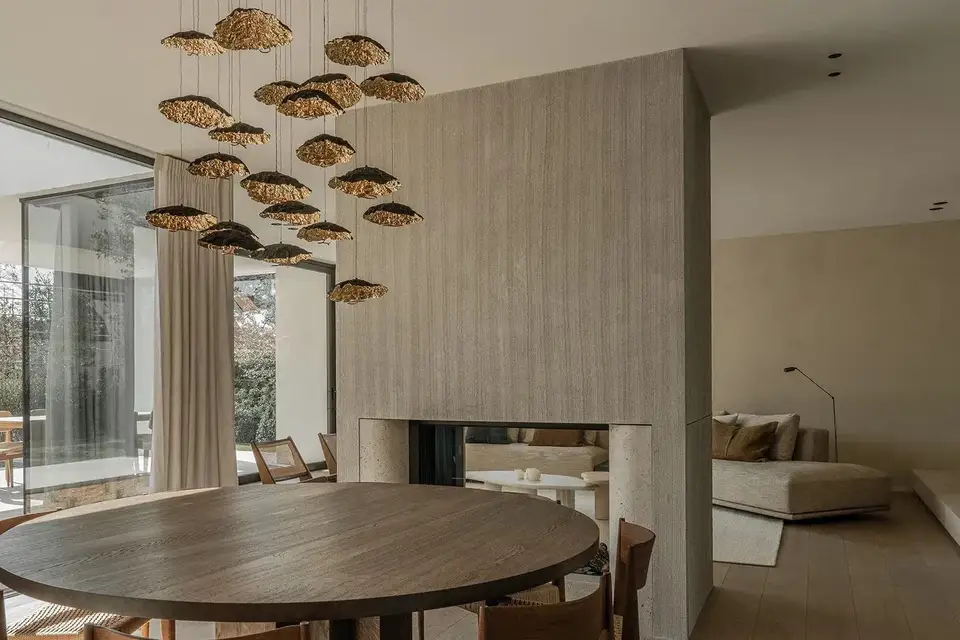 Een ronde houten eettafel naast een doorkijkhaard van travertin, onder een wolk van bronskleurige schijven aan dunne kabels