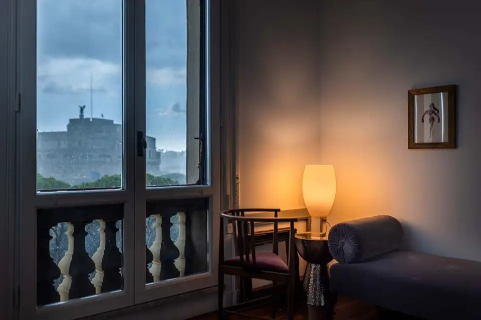 Een glazen tafellamp brandt in de schemer naast een hoog raam met een stenen balustrade