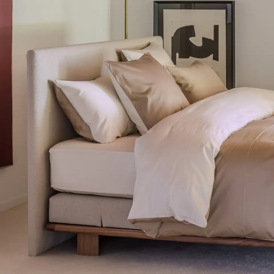 Een bed op een houten onderstel met champagnekleurig satijnen beddengoed, een messing wandlamp en abstracte kunst aan de wand