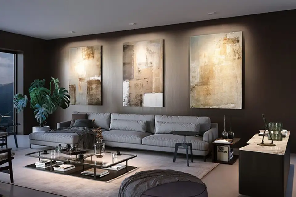 Plafondspots strijken licht over drie grote schilderijen boven een grijze bank in een donkere woonkamer