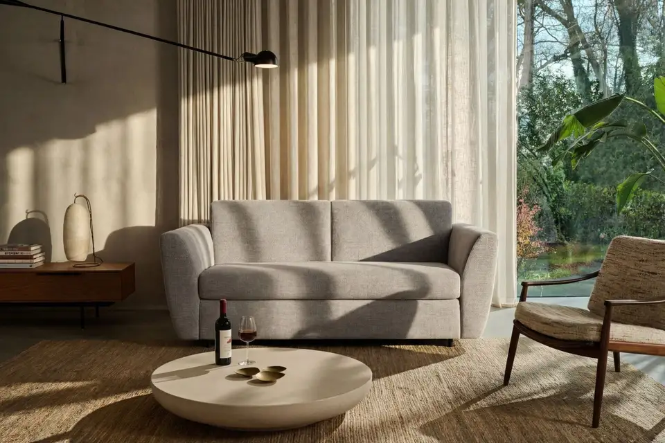 De slaapbank Vinnie in een lichte stof in warm tegenlicht, met linnen vitrages, een ronde salontafel en een houten fauteuil