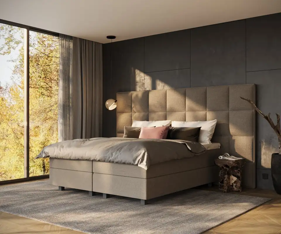 Een taupe boxspring met een hoog hoofdbord van gestoffeerde vlakken tegen een donkere wand, met een glazen hanglamp en avondzon door een groot raam