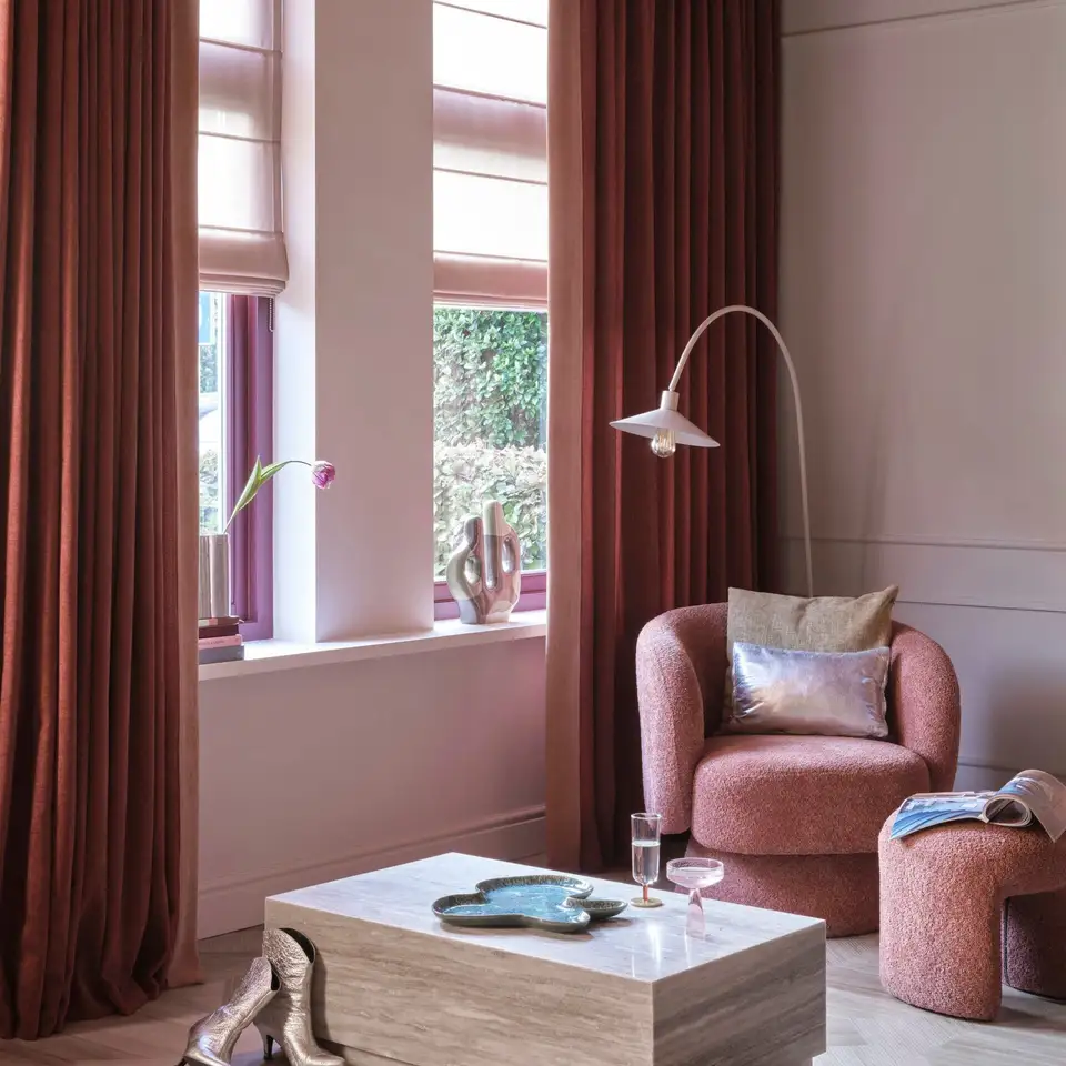 Diep rode gordijnen en roze vouwgordijnen voor hoge ramen, met een roze fauteuil en een travertin salontafel