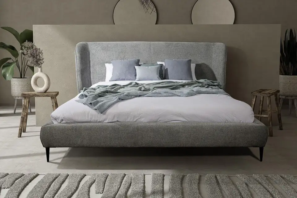 Het bed Palm met een gebogen, grijs gestoffeerd hoofdbord en slanke zwarte poten, tegen een beige wand met ronde wandschijven