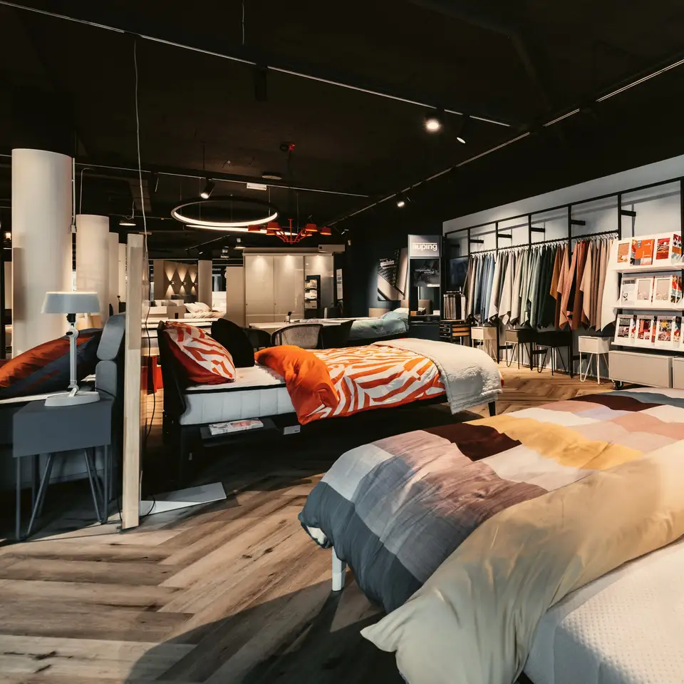 De bedvloer van onze showroom: een opgemaakt bed met een oranje dekbed, een lichtring aan het plafond en een rek met stoffen, op een visgraatvloer