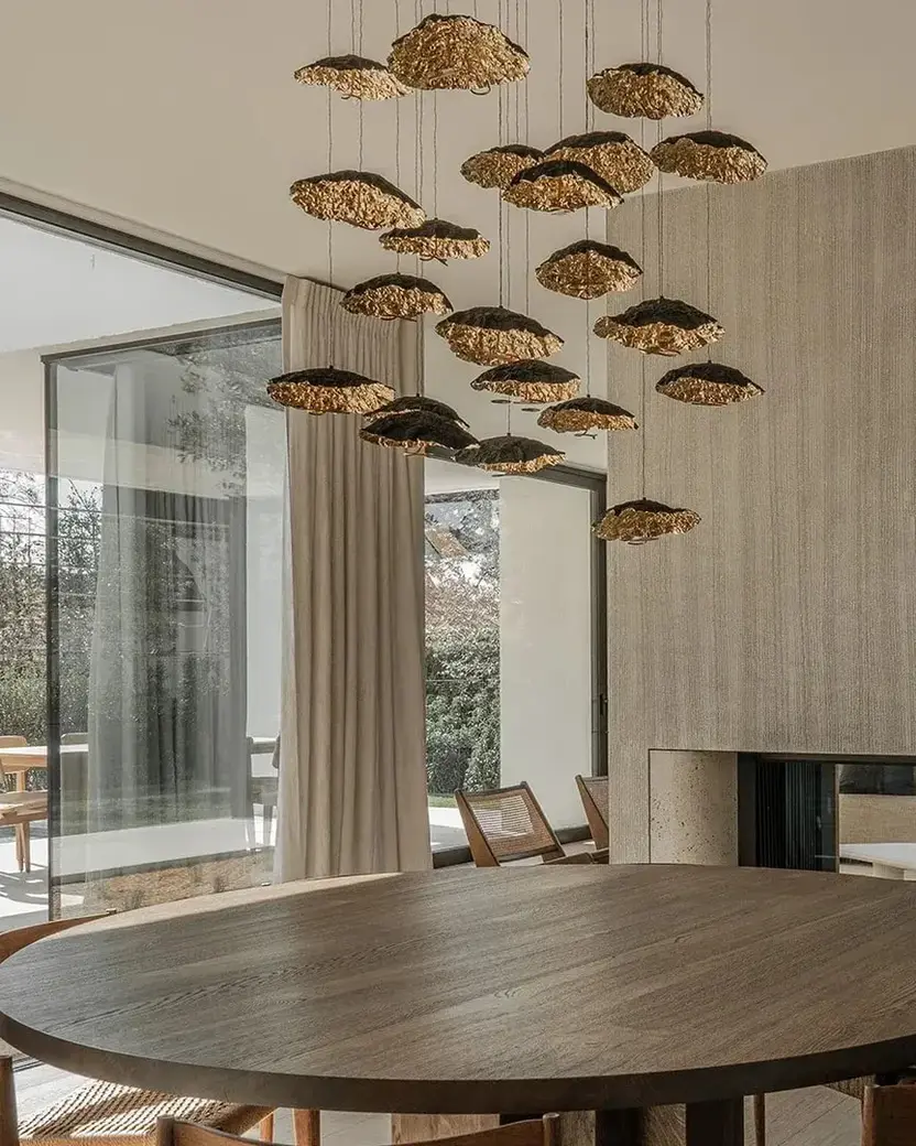 Een ronde eiken eettafel onder een wolk van bronzen bladvormige hanglampen, naast een doorkijkhaard