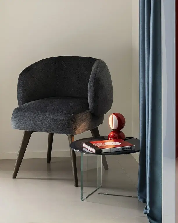 Een kleine rode tafellamp op een bijzettafel naast een grijze fauteuil