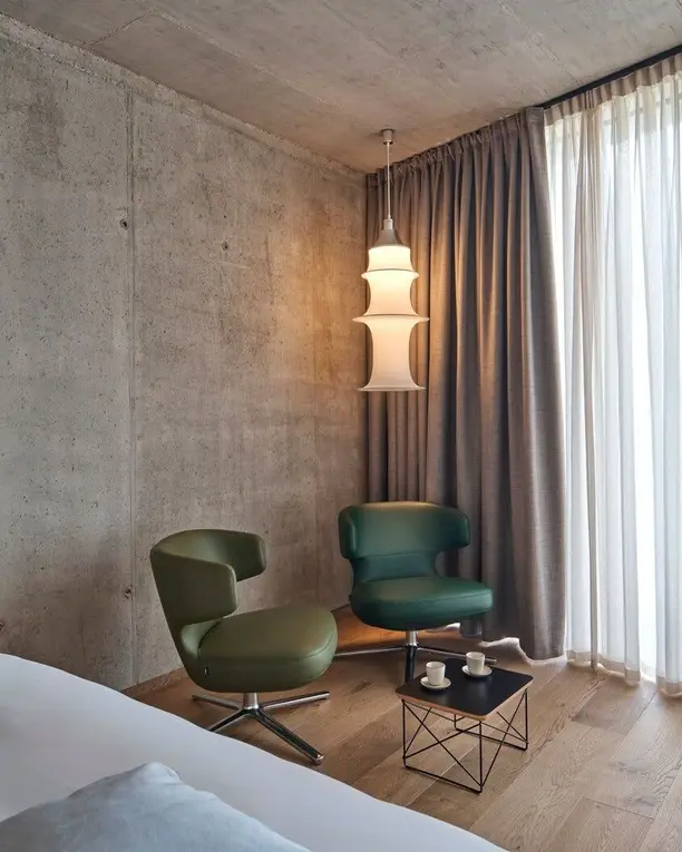 Een witte hanglamp in een slaapkamer met betonnen wanden en twee groene fauteuils