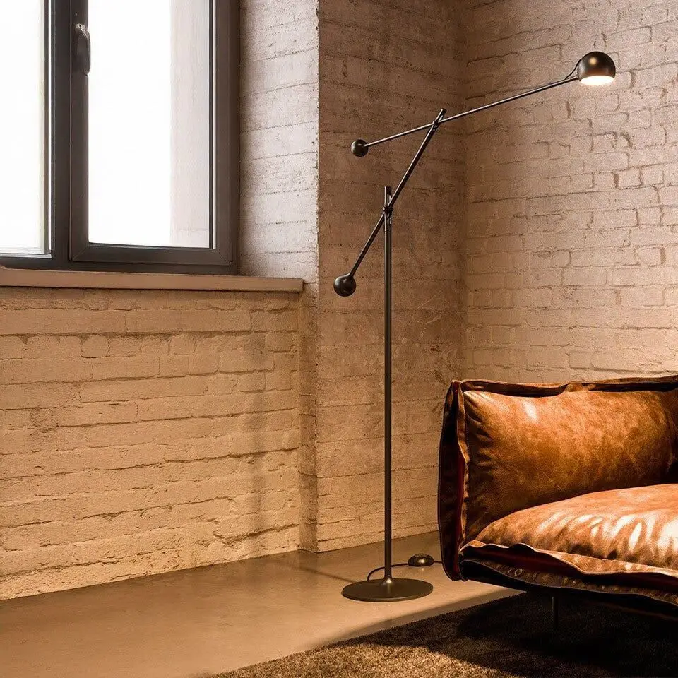 Een zwarte vloerlamp met een lange arm en een kleine ronde kop naast een leren bank bij een raam