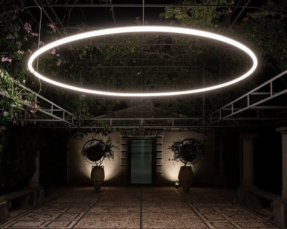 Een grote cirkel van licht hangt in een donker tuinpaviljoen met zuilen en planten