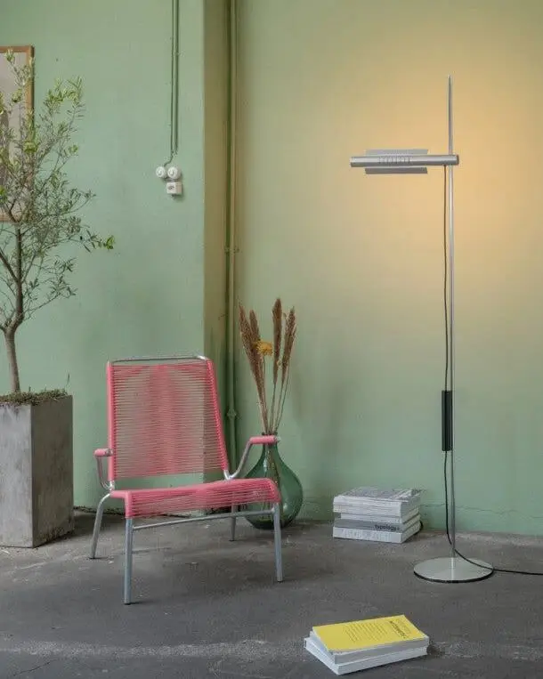 Een zilveren vloerlamp met een platte kop naast een roze stoel tegen een groene wand