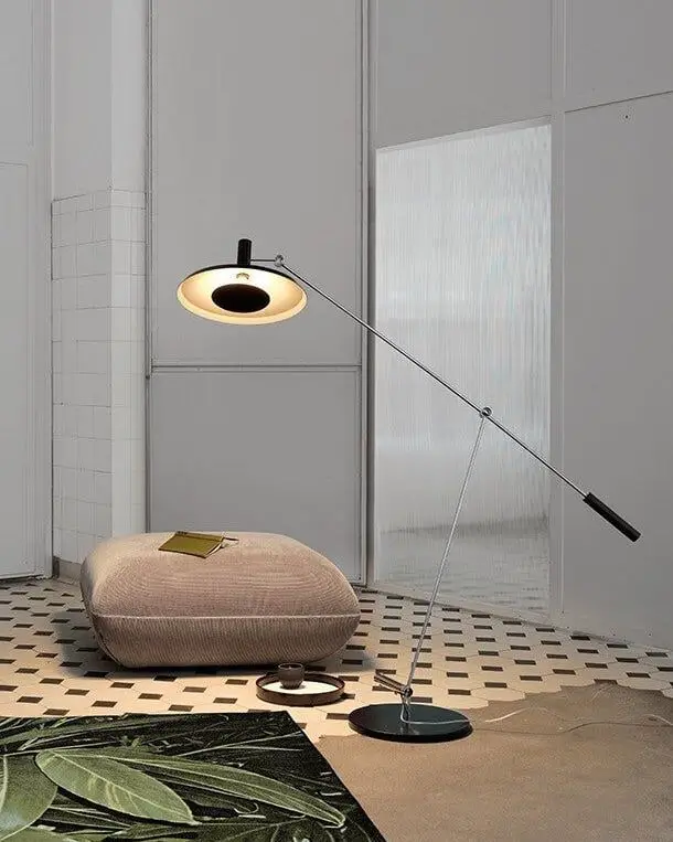 Een vloerlamp met een lange arm en een ronde reflector naast een poef op een tegelvloer