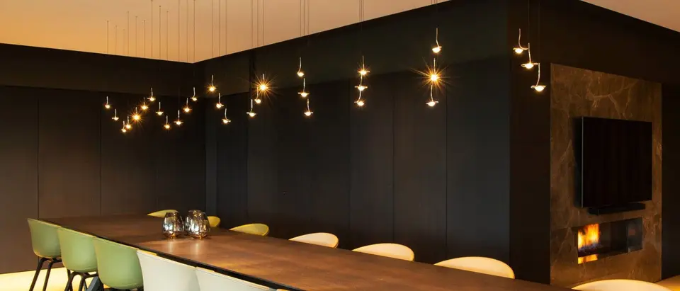 Een lange notenhouten tafel met lichte stoelen in een donkere zaal, onder tientallen kleine lichtpunten
