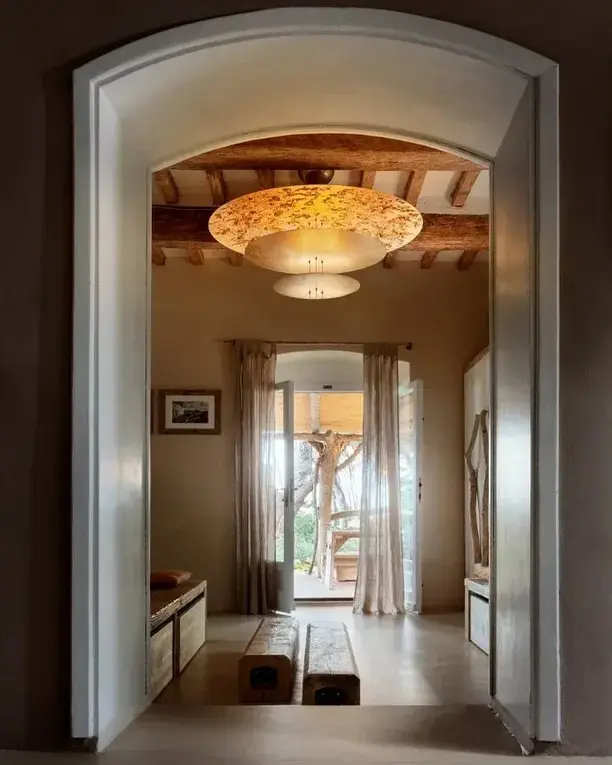 Doorkijk door een witte boog naar een goudkleurige plafondlamp in een kamer met houten balken