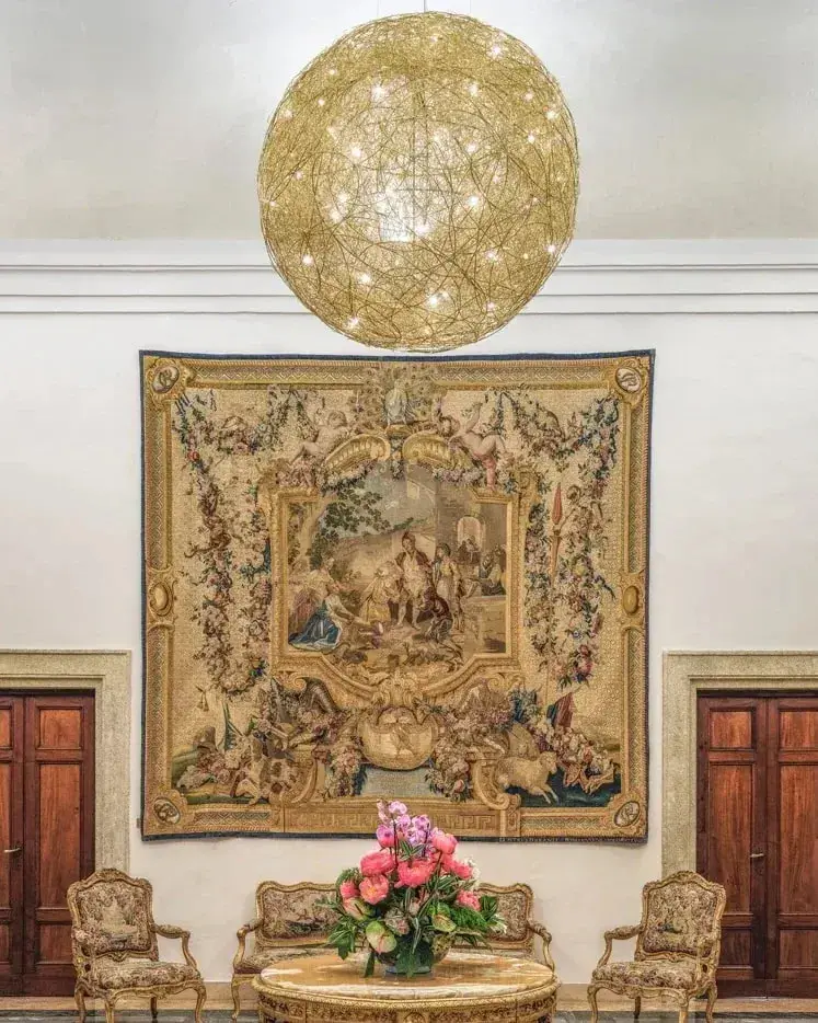 Een grote bol van gouden draad hangt in een zaal met een wandtapijt en klassieke stoelen