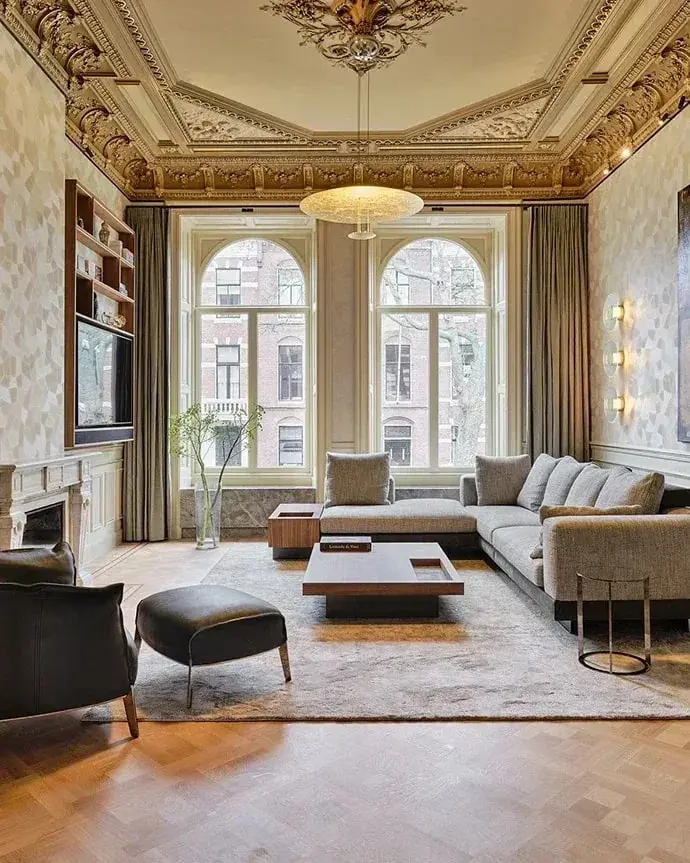 Een gouden plafondlamp in een hoge kamer met stucplafond, boogramen en een lichte hoekbank