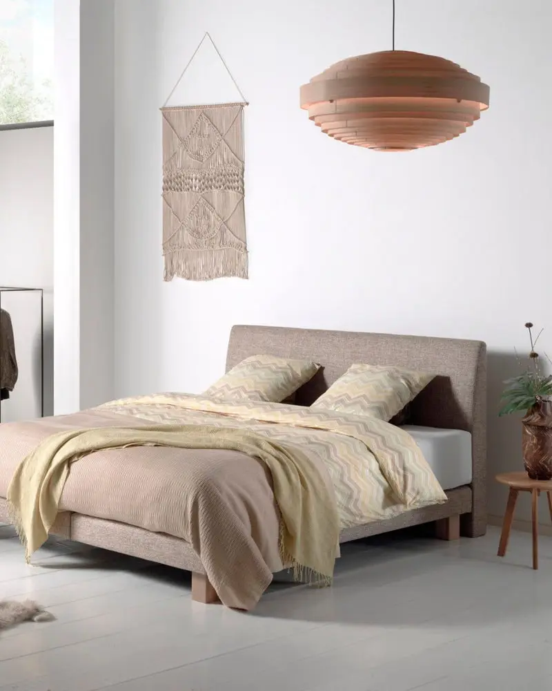 Een Balance-boxspring met een beige gestoffeerd hoofdbord in een lichte kamer met een koperen hanglamp en een schapenvacht