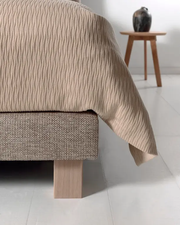 Het voeteneinde van een boxspring op een houten blokpoot, met een golvend dekbed en een krukje met een vaas