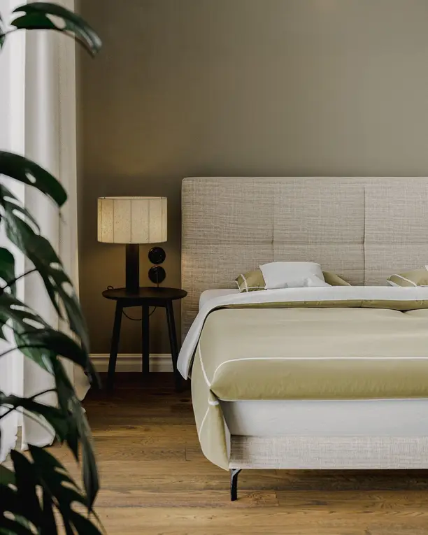 Het bed met geruit hoofdbord van opzij, met een tafellamp en een plant tegen de olijfgroene wand