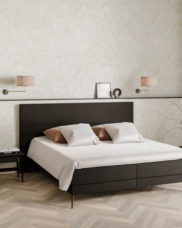 Een donkere boxspring met een hoog hoofdbord en roze kussens op een lichte visgraatvloer