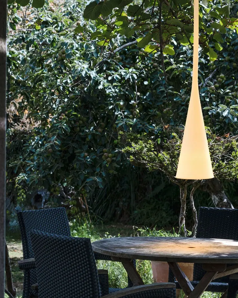 Een witte kegelvormige lamp hangt boven een tuintafel, gezien vanuit een openslaande deur