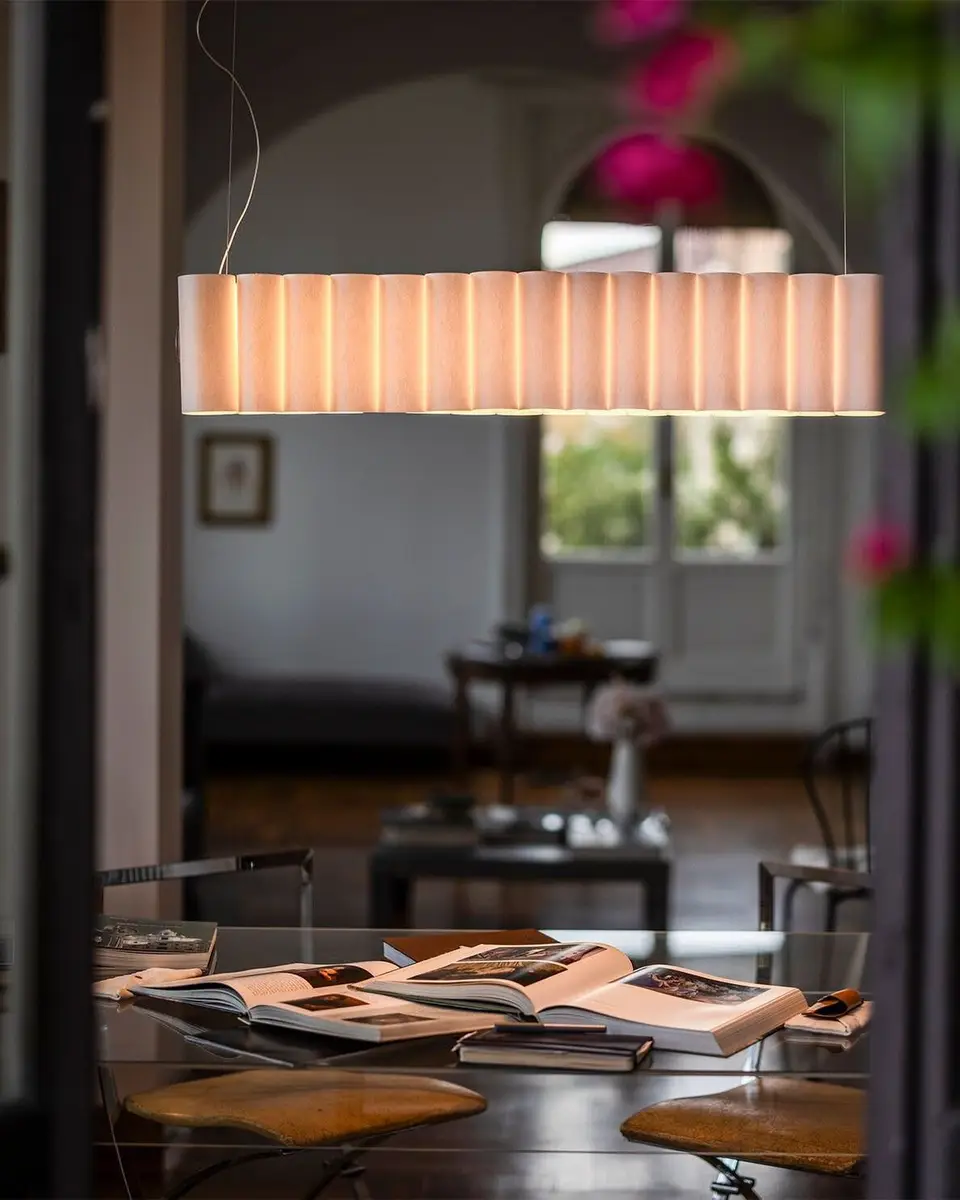 Een lange geplooide hanglamp boven een eettafel, gezien door een deuropening met roze bloemen