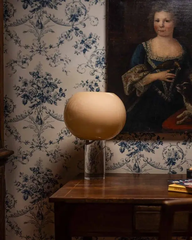 Een ronde glazen tafellamp op een bureau voor blauw bloemenbehang en een portret