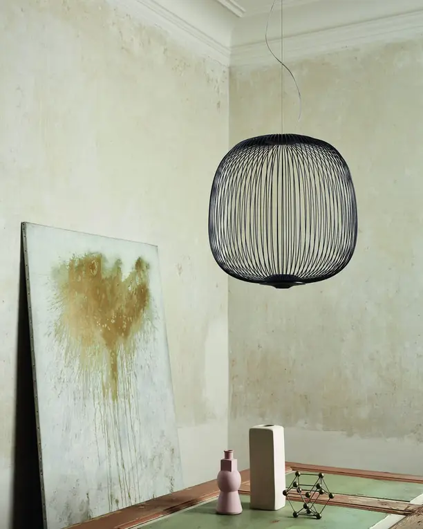 Een donkere hanglamp van dunne spaken in een lichte hoek met een schilderij tegen de wand