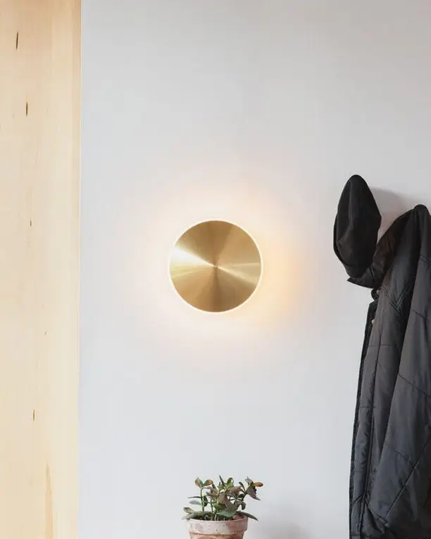 Een ronde messing wandlamp naast een kapstok met een jas