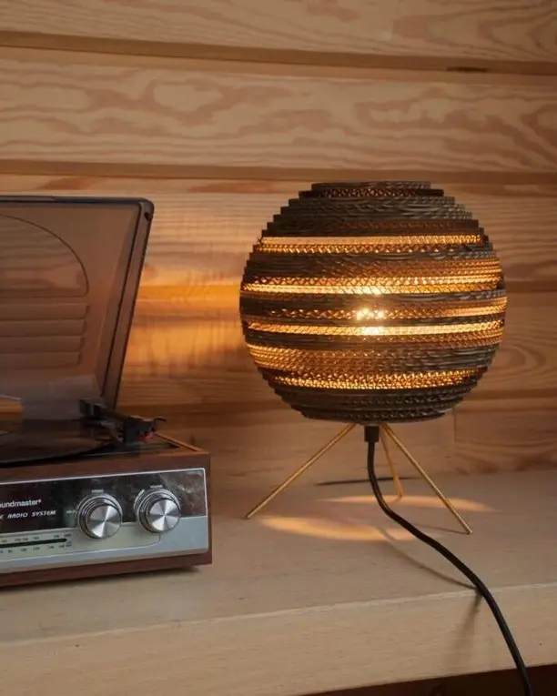 Een bolle lamp van karton op een driepoot naast een platenspeler, tegen een houten wand