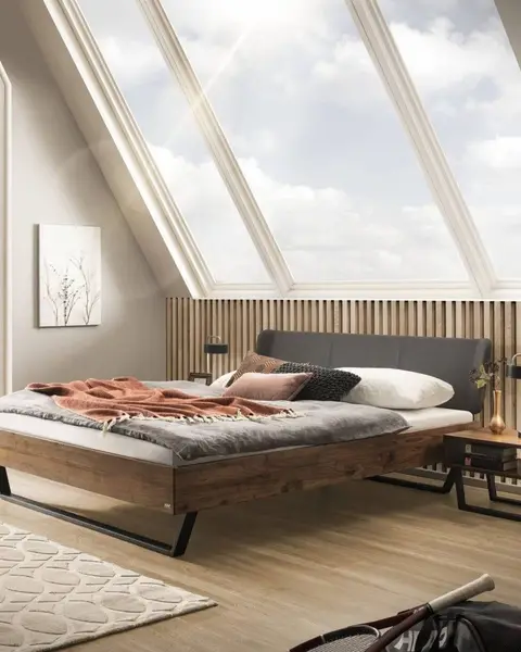 Een houten bed met een donker hoofdbord en een roestrode plaid onder schuine dakramen, op een lichte houten vloer