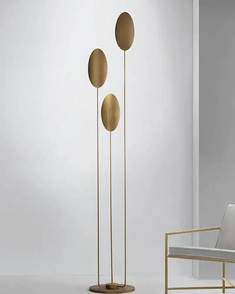 Een staande lamp met drie gouden schijven op dunne stelen naast een stoel