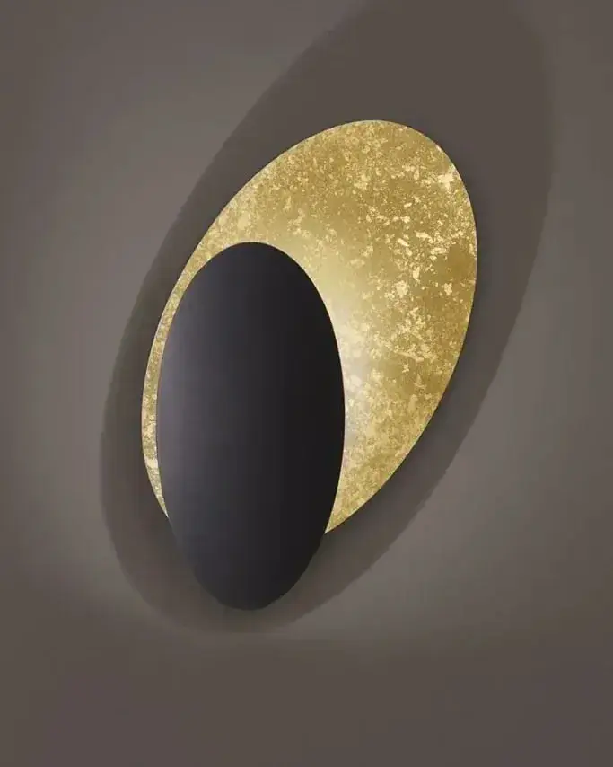 Een wandlamp met een schijf in bladgoud en een zwarte schijf op een grijze wand