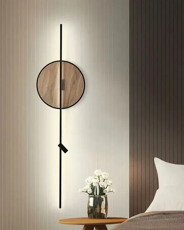 Een houten schijf aan een verticale lichtlijn op een wand naast een bed en een bijzettafel met bloemen