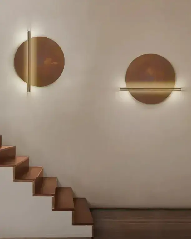 Twee koperkleurige schijven met een lichtlijn aan een lichte wand boven een houten trap