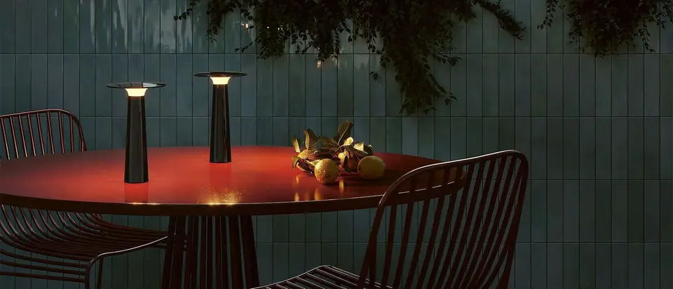 Twee draagbare lampen op een rode tuintafel bij avond, tegen een groen betegelde muur met hangende planten