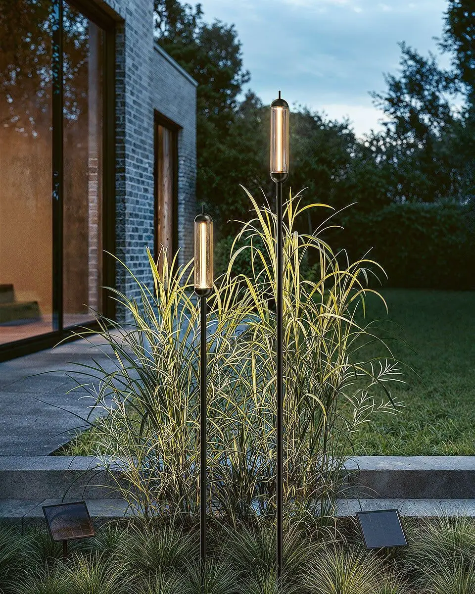 Twee slanke tuinlampen op een pen tussen hoge grassen bij een bakstenen huis, bij schemer