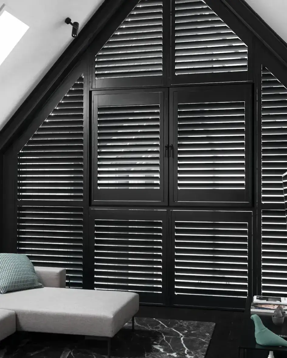 Zwarte shutters in een driehoekig raam onder de nok, met een lichte hoekbank ervoor