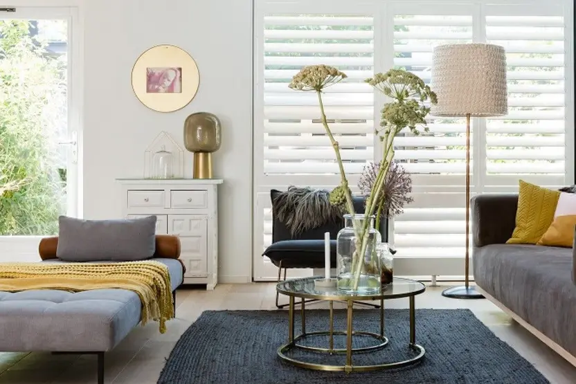 Witte shutters voor een hoog raam in een lichte woonkamer met een glazen salontafel