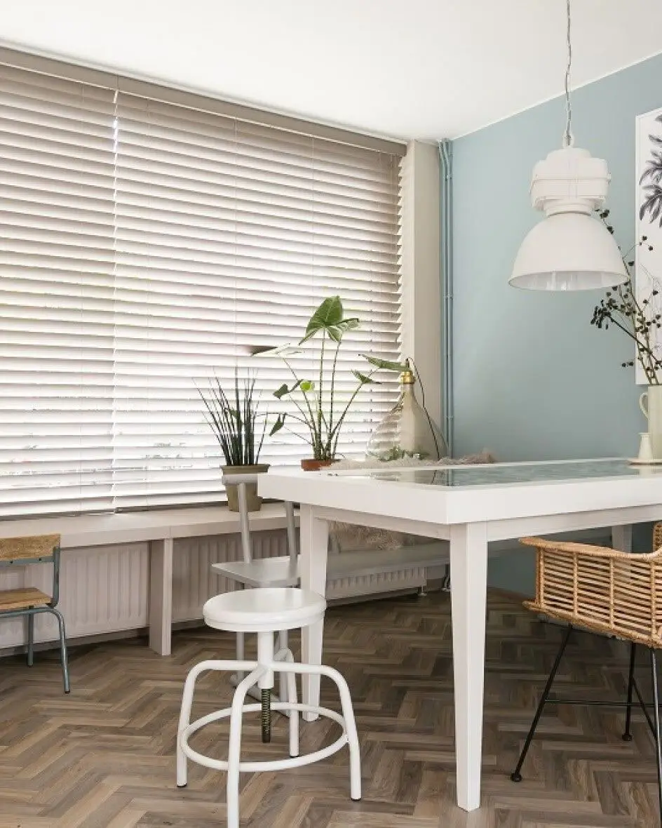 Houten jaloezieën boven een witte tafel met rotan stoelen, voor een lichtblauwe wand