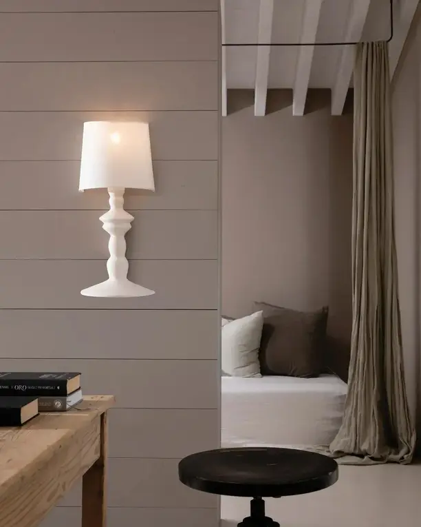 Een witte wandlamp met kap naast een doorgang naar een zithoek met gordijnen