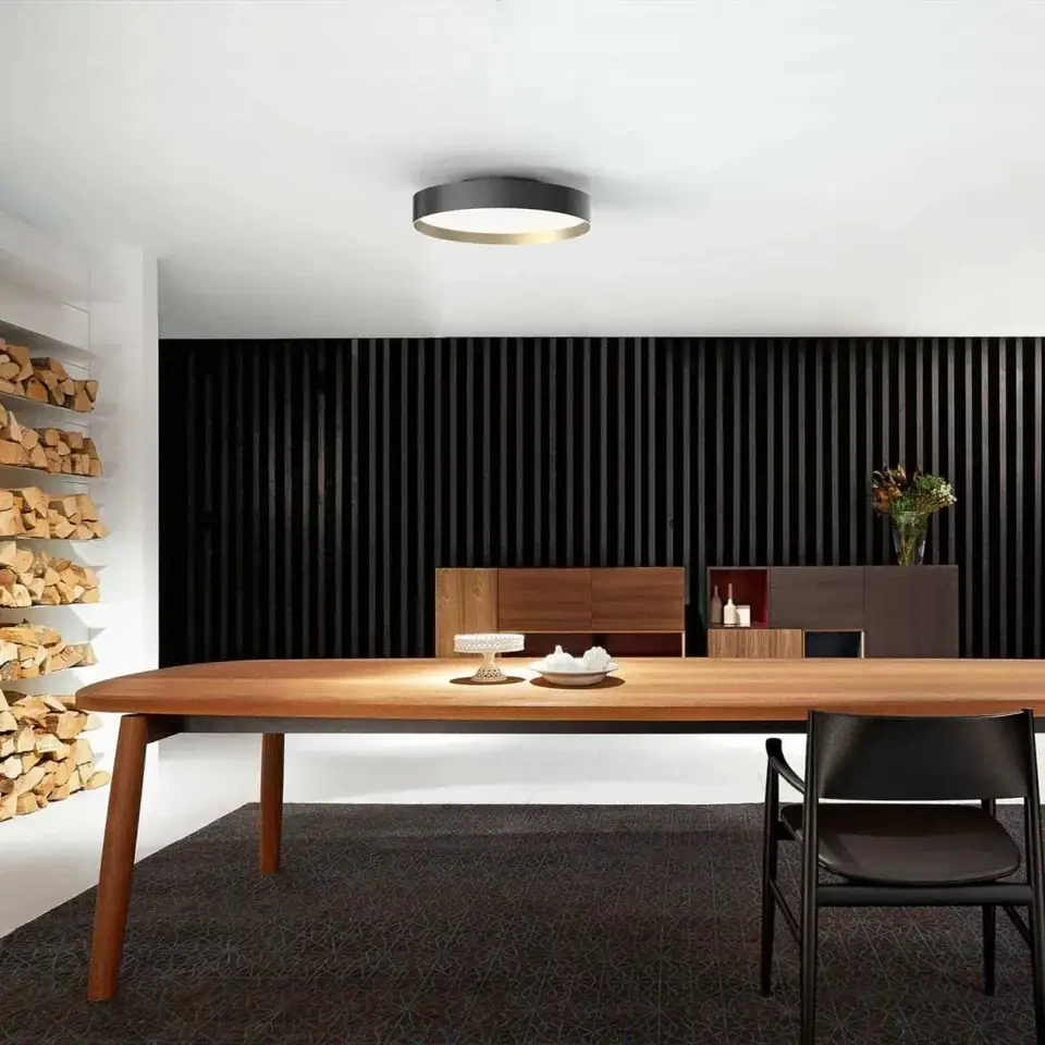 Een ronde zwarte plafondlamp met een gouden rand boven een lange notenhouten tafel, voor een zwarte latjeswand met gestapeld haardhout
