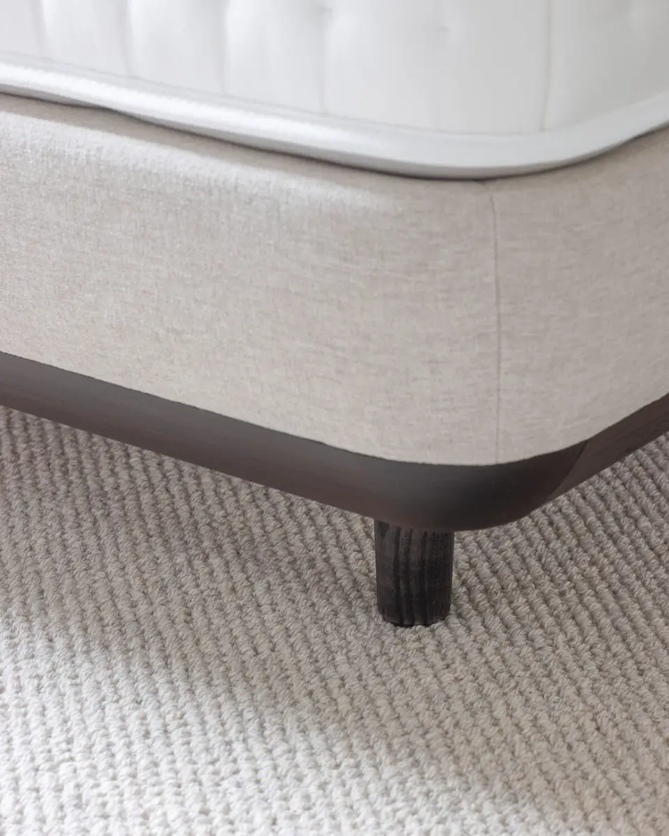 Hoek van een getufte matras op een boxspring in lichte linnen stof, met een donkere plint en poot op een wollen kleed