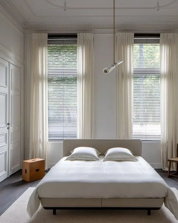 Een lichte boxspring midden in een hoge kamer van een herenhuis, tussen twee ramen met vitrages