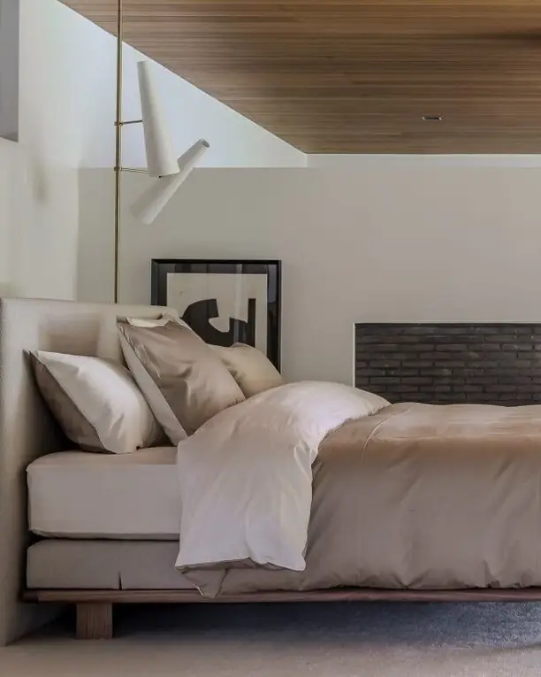 Een laag bed met champagnekleurig satijn naast een open haard van zwarte baksteen, onder een houten plafond